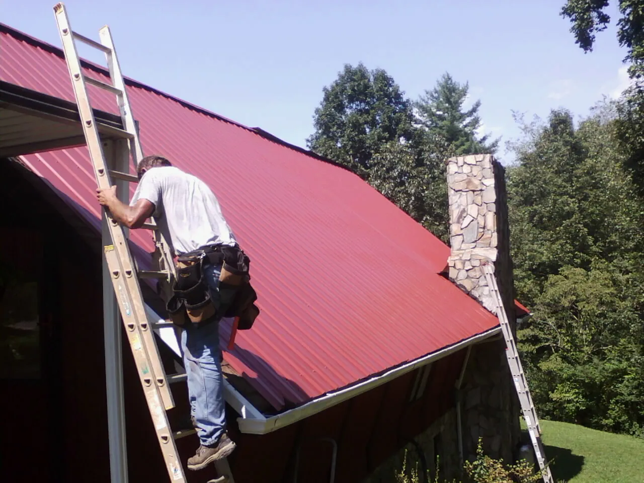 Roofer on a ladder beside a red metal roof and stone chimney — Soffit & Fascia Repair by Roofing Brothers in Murdock